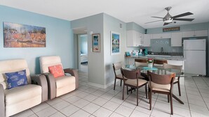A living room with a sofa, a dining table with chairs, a kitchen area with cabinets and a refrigerator, and a ceiling fan. - Oceanfront Condo with Rocket Launch Views - Steps to Beach (Cocoa Beach)