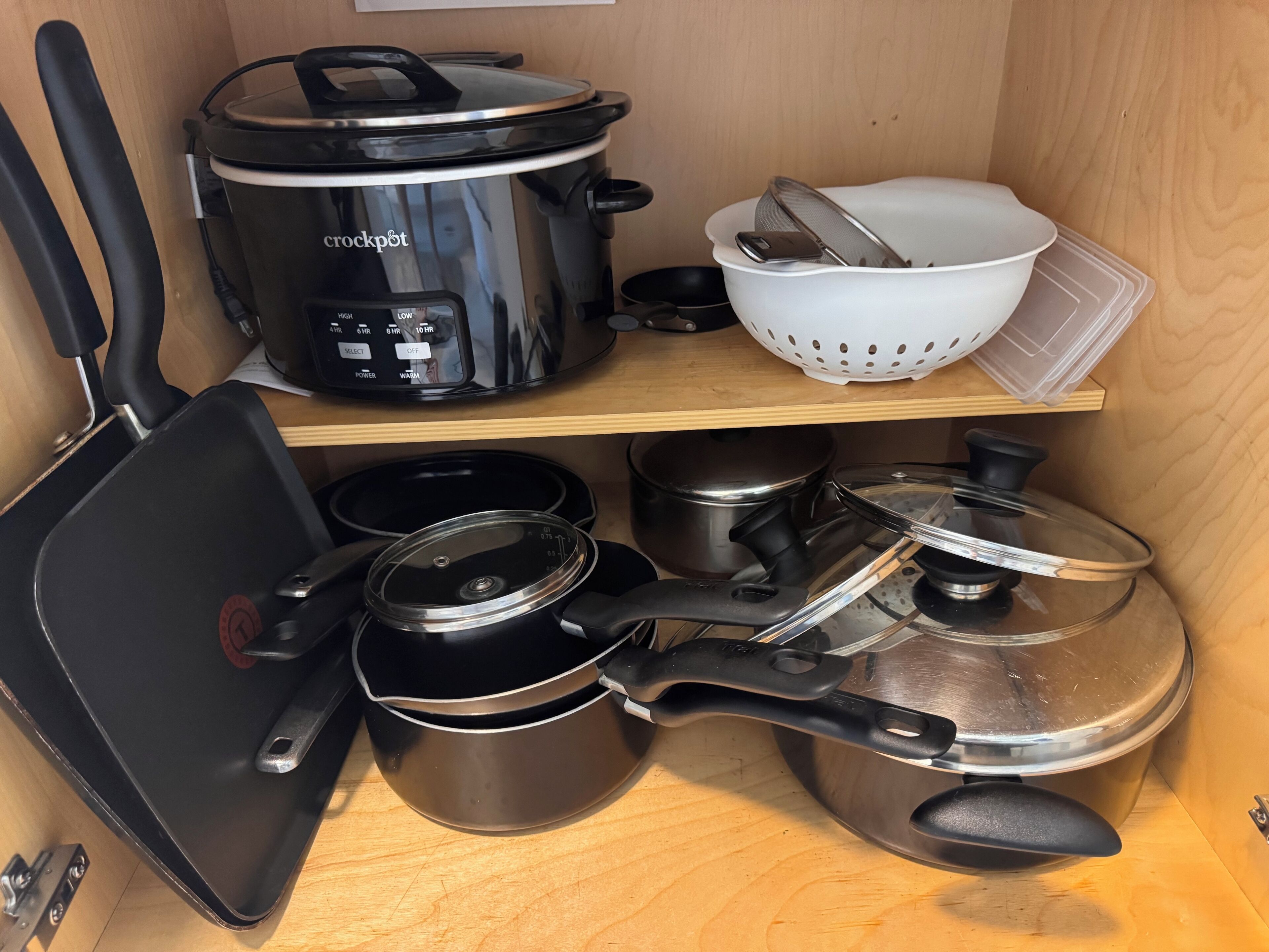 A kitchen with a crockpot, pots, a frying pan, and a colander on shelves.