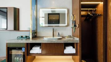 Un baño moderno con lavabo, espejo, toallero y un refrigerador pequeño.