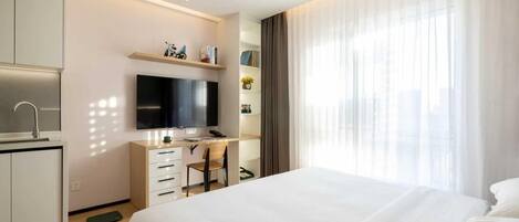 A modern hotel room with a bed, a desk with a chair, a TV, and a window with curtains.
