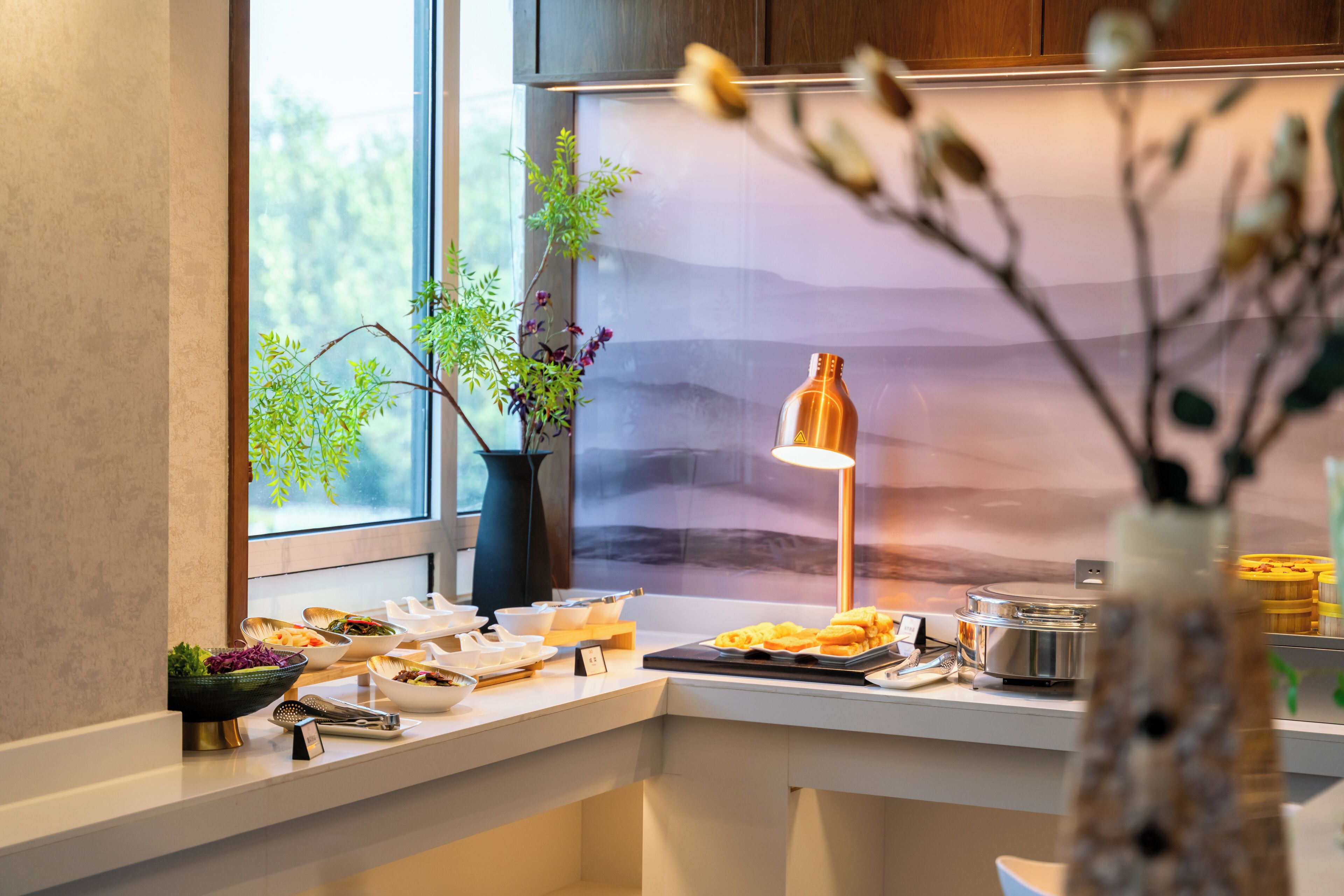 A buffet setup with various food items, a vase with flowers, and a lamp.
