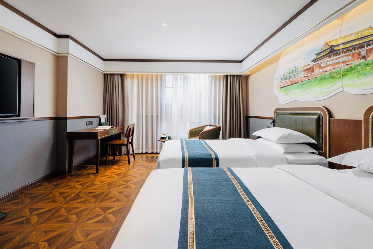 A hotel room with two beds, a desk, a chair, a television, and a large window with curtains.