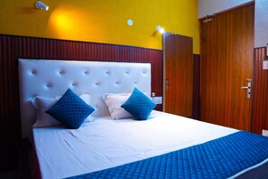 Quarto de hotel com uma cama grande, dois travesseiros azuis, um edredom branco e um guarda-roupa de madeira.