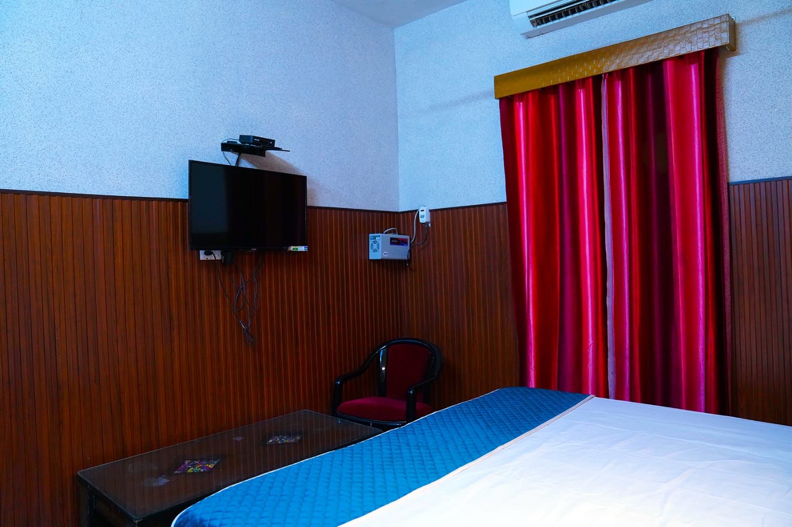 Habitación de hotel con cama, televisión instalada en la pared, escritorio con silla y ventana con cortinas rojas.