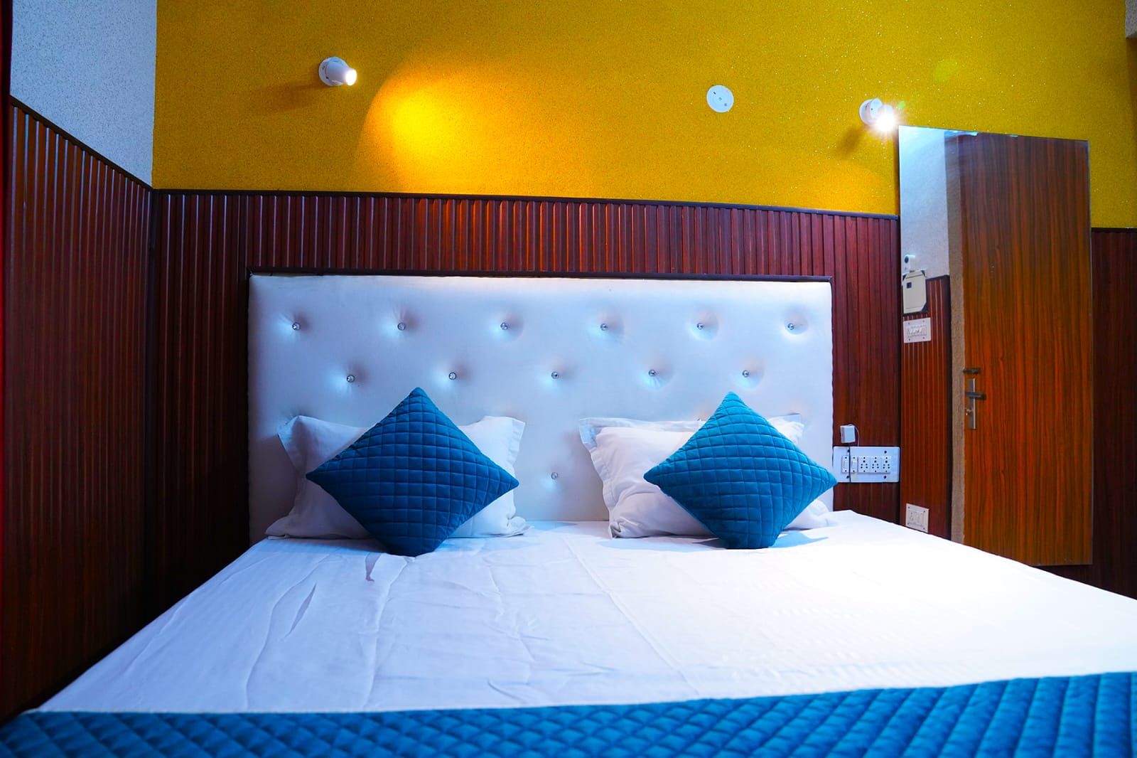 A hotel room with a large bed, two blue pillows, a white headboard, and wooden paneling on the walls.