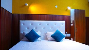 A hotel room with a large bed, two blue pillows, a white headboard, and wooden paneling on the walls. - Apna Niwas (Haroli)