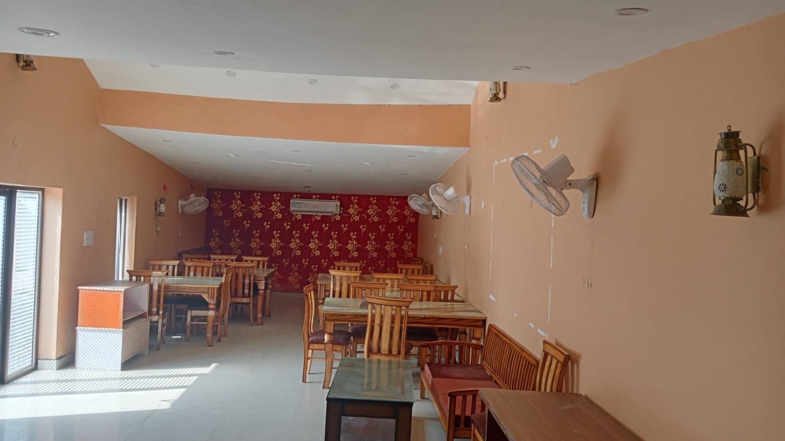 A dining area with wooden tables and chairs, a red patterned wall, and ceiling fans.