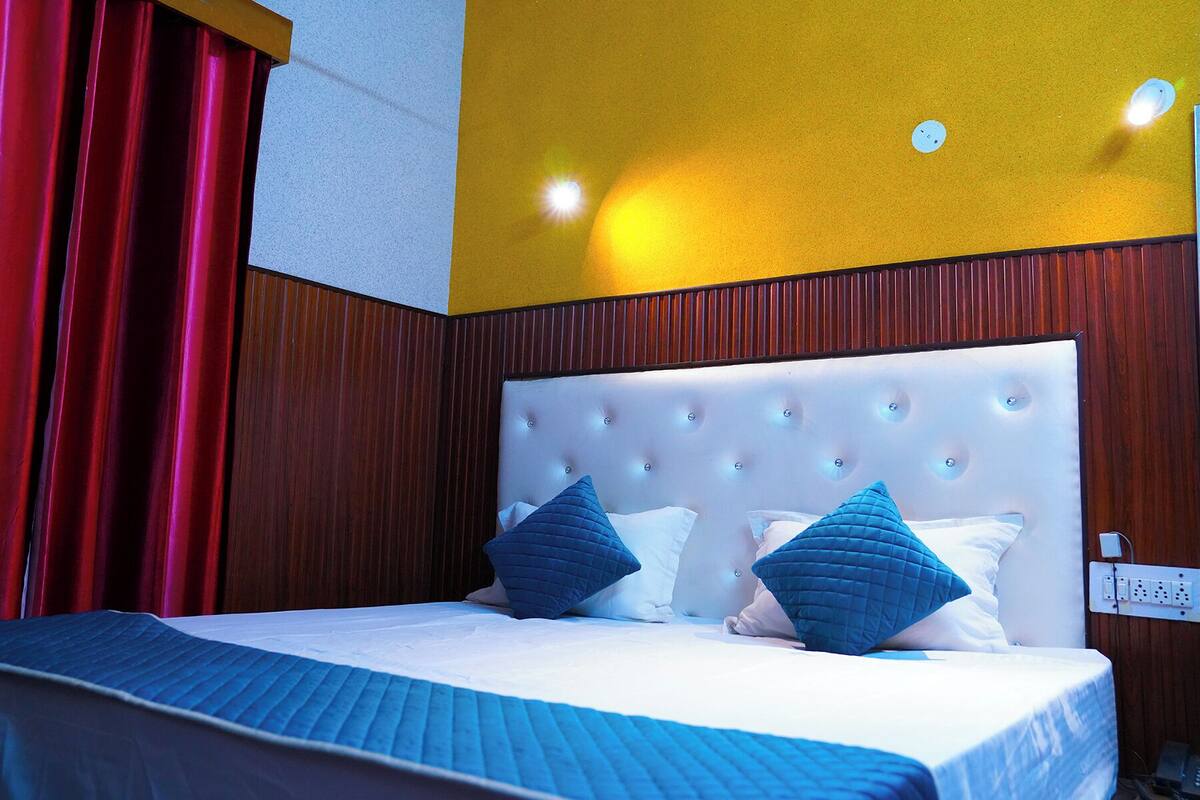 Quarto de hotel com cabeceira estofada, cama com roupa de cama branca e azul, e cortinas vermelhas.