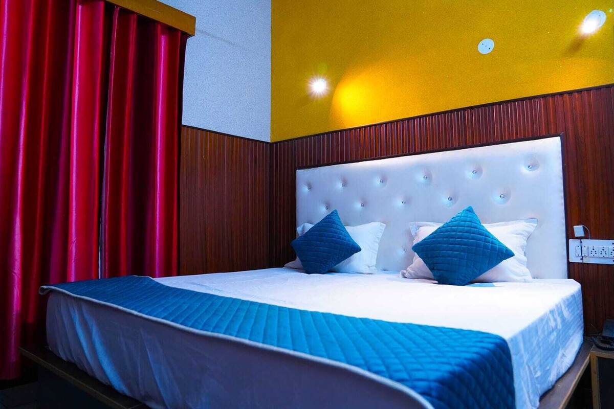 Quarto de hotel com uma cama grande, cortinas vermelhas, cabeceira branca e uma parede amarela.