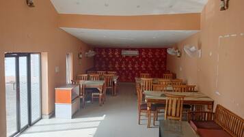 A dining area with wooden tables and chairs, a red patterned wall, and a window with blinds.
