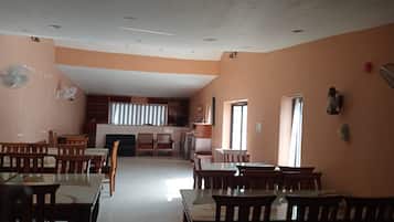 Uma sala de jantar com várias mesas e cadeiras, área de cozinha com armários e eletrodomésticos, e uma janela grande que traz luz natural.
