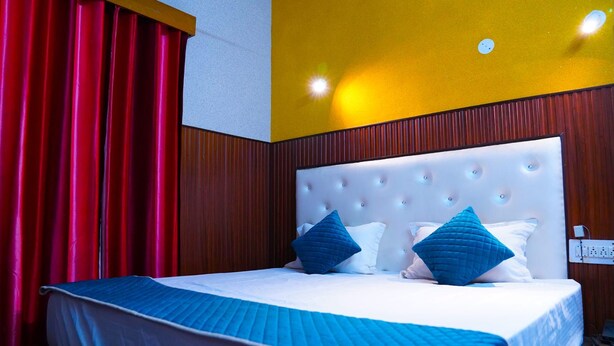 A hotel room with a large bed, red curtains, a white headboard, and a yellow accent wall. - Apna Niwas (Haroli)