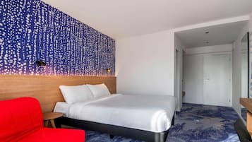 A hotel room with a bed, a red chair, a wooden headboard, and a wall with a blue and white pattern.