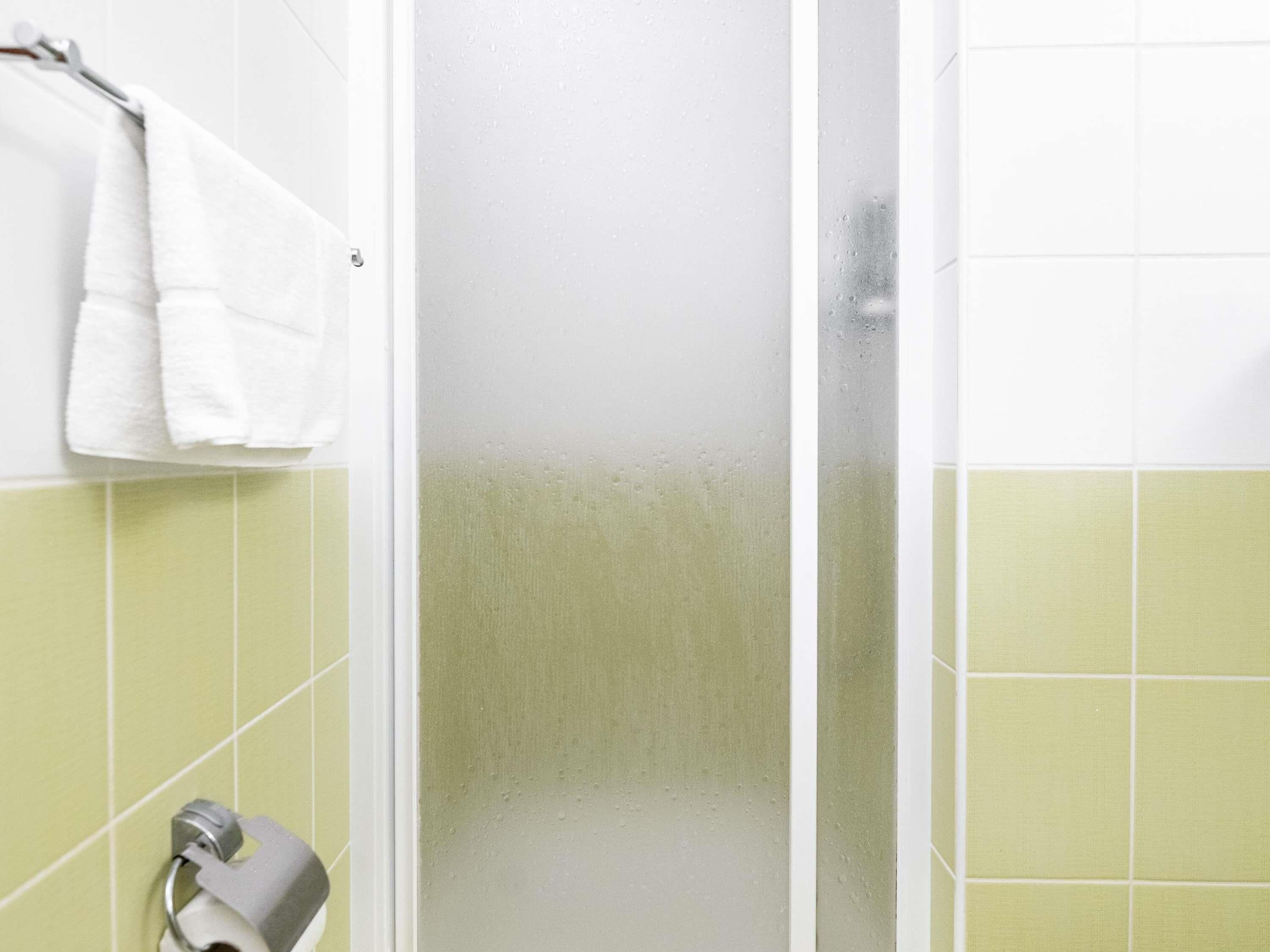 A bathroom with a glass shower door, a towel rack with a white towel, and green tiled walls.