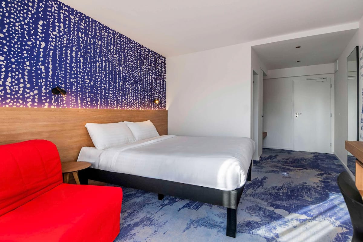 A hotel room with a bed, a red chair, a wooden headboard, and a wall with a blue and white pattern.