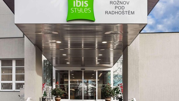 Entrada de um edifício moderno com uma placa verde que diz “ibis Styles” e “ROŽNOV POD RADHOŠTĚM”.
