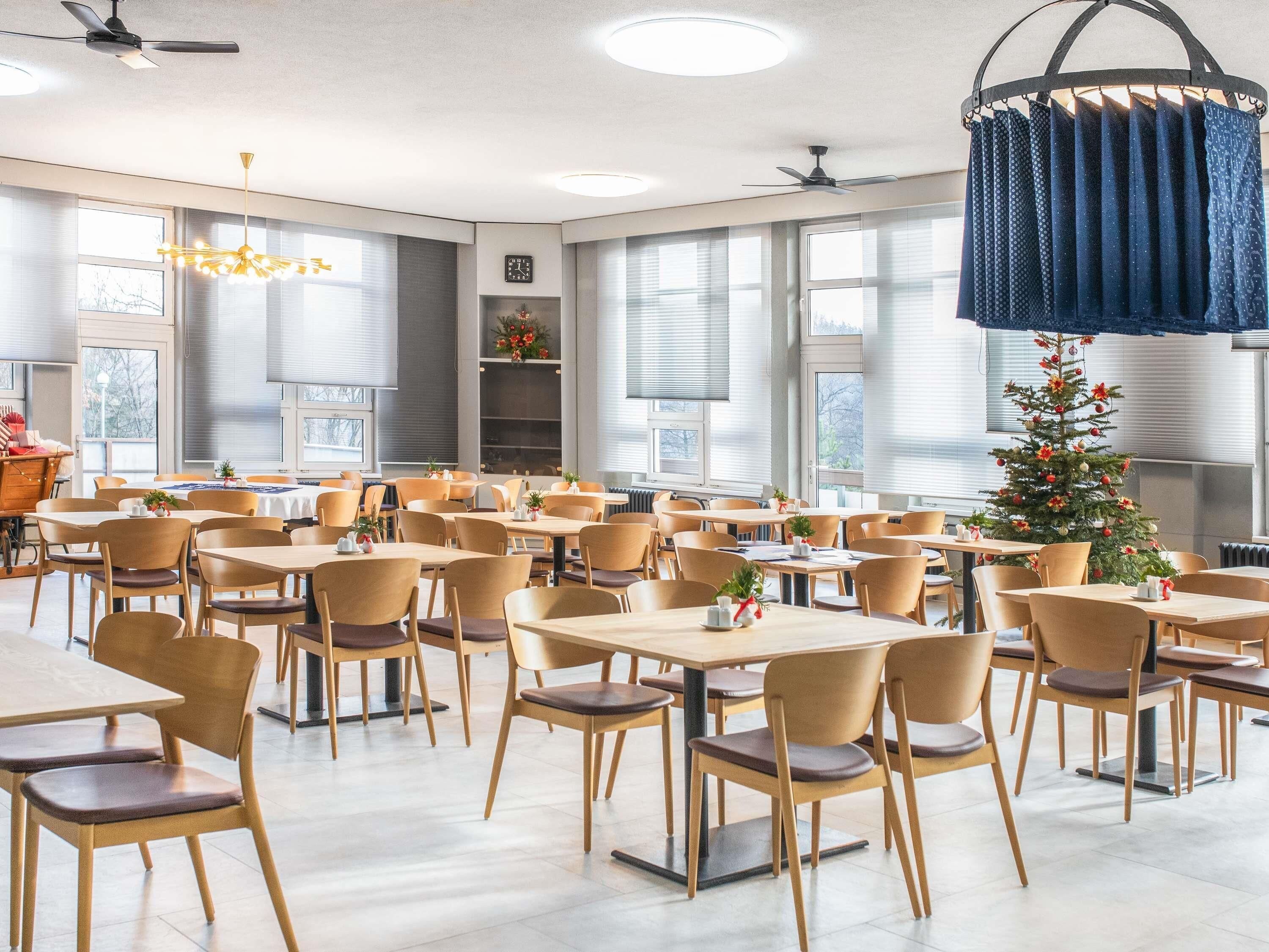 Un comedor amplio con varias mesas y sillas, ventanas grandes, ventiladores de techo y un árbol decorativo navideño.