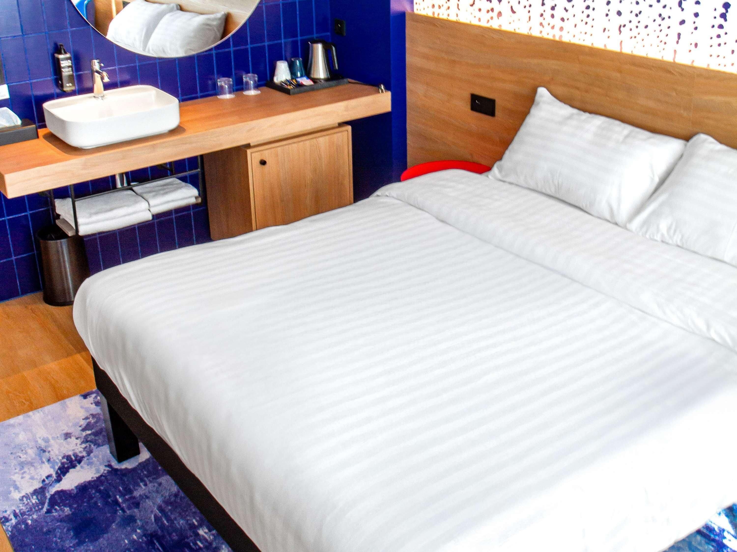 Una habitación de hotel moderna con una cama grande, cabecera de madera, lavamanos integrado y un espejo.