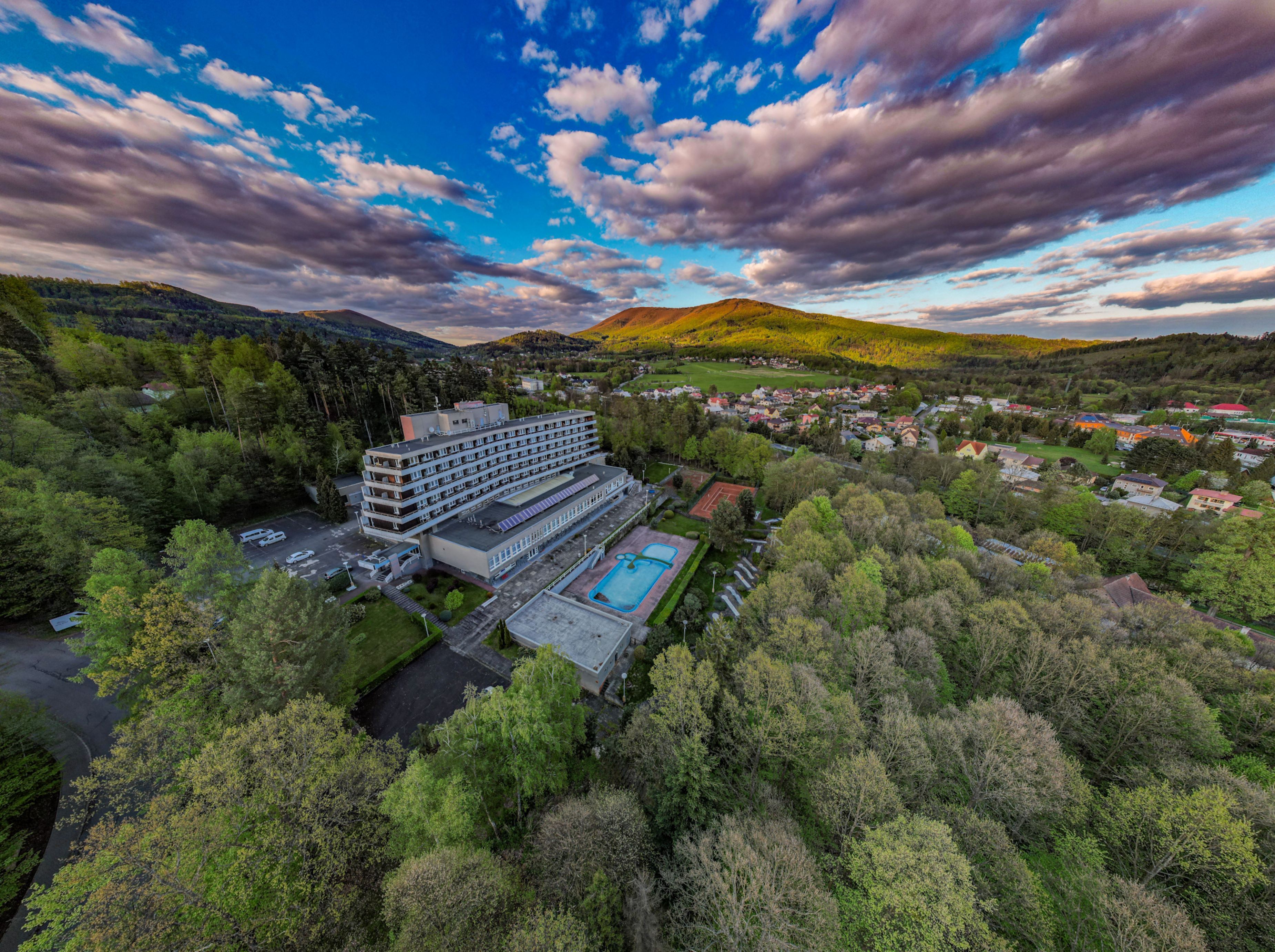 An aerial view of a hotel with multiple floors, a swimming pool, and a parking area surrounded by dense forest.