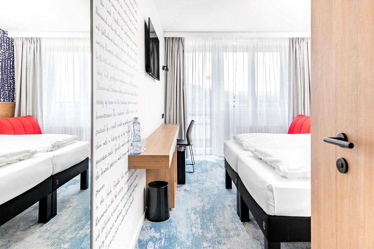 A hotel room with two beds, a wooden bench, a small table, a TV mounted on the wall, and a large window with sheer curtains.