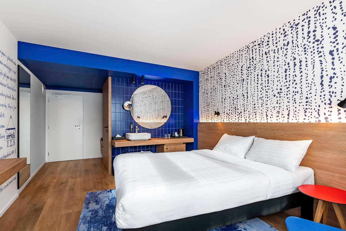 A modern bedroom with a large bed, a round mirror, a wooden nightstand, and a red side table.