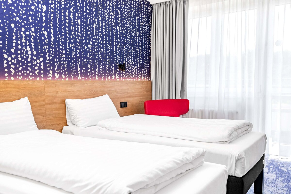 Ein Hotelzimmer mit zwei Betten, einem Holzkopfende, einem roten Stuhl und einem Fenster mit transparenten Vorhängen.