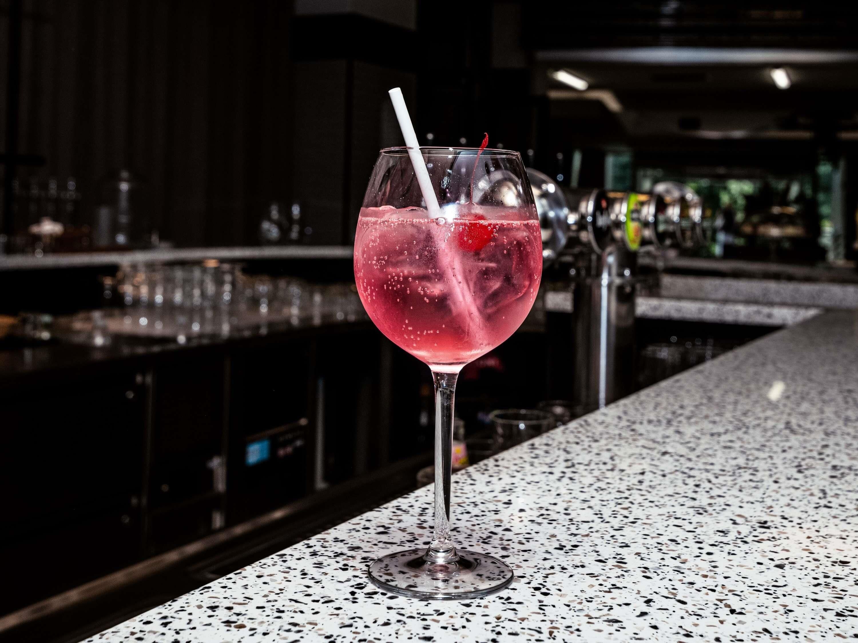 Un bar con un vaso de cóctel rosado, hielo y una pajita, sobre una encimera con motas.