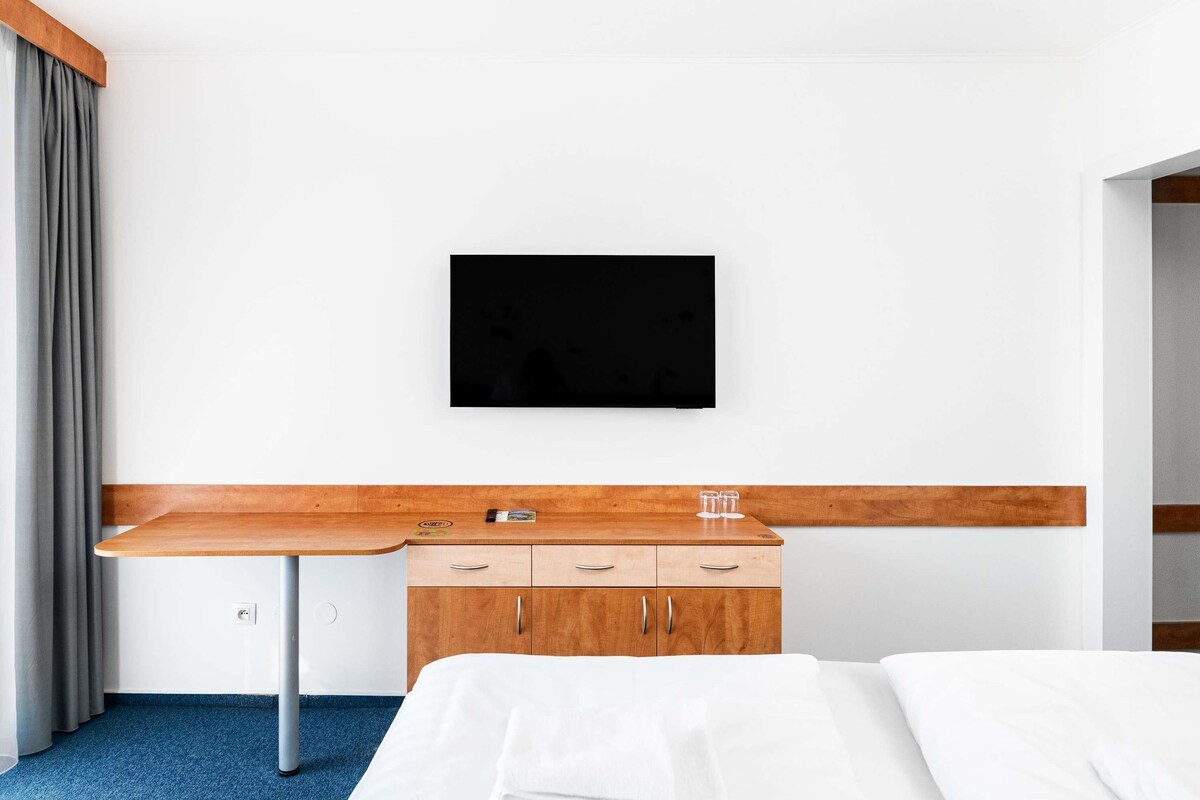 A hotel room with a bed, a wooden desk, a television mounted on the wall, and a wardrobe.