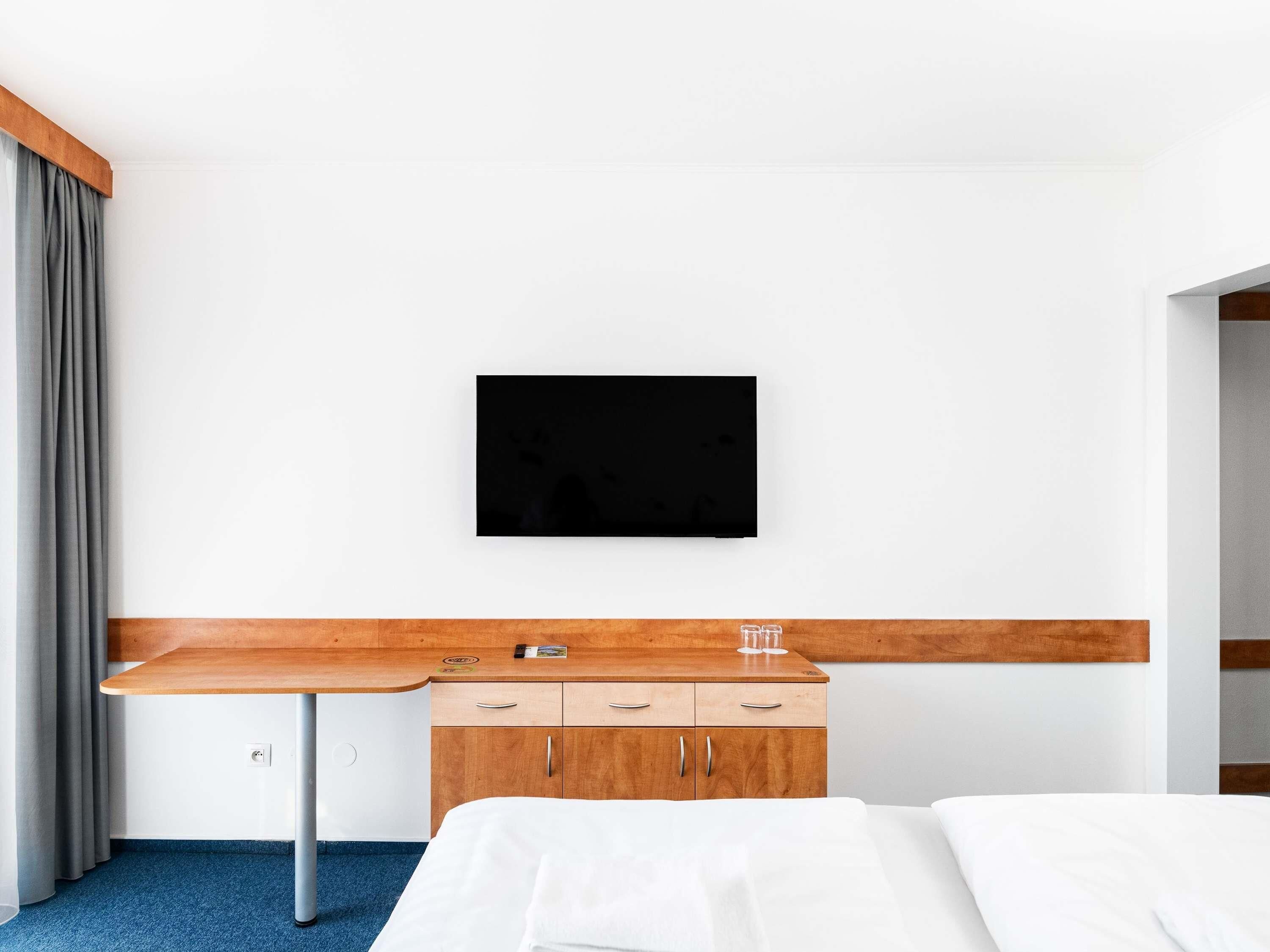 Ein Hotelzimmer mit einem Bett, einem Holztisch, einem an der Wand befestigten Fernseher und einem Kleiderschrank.