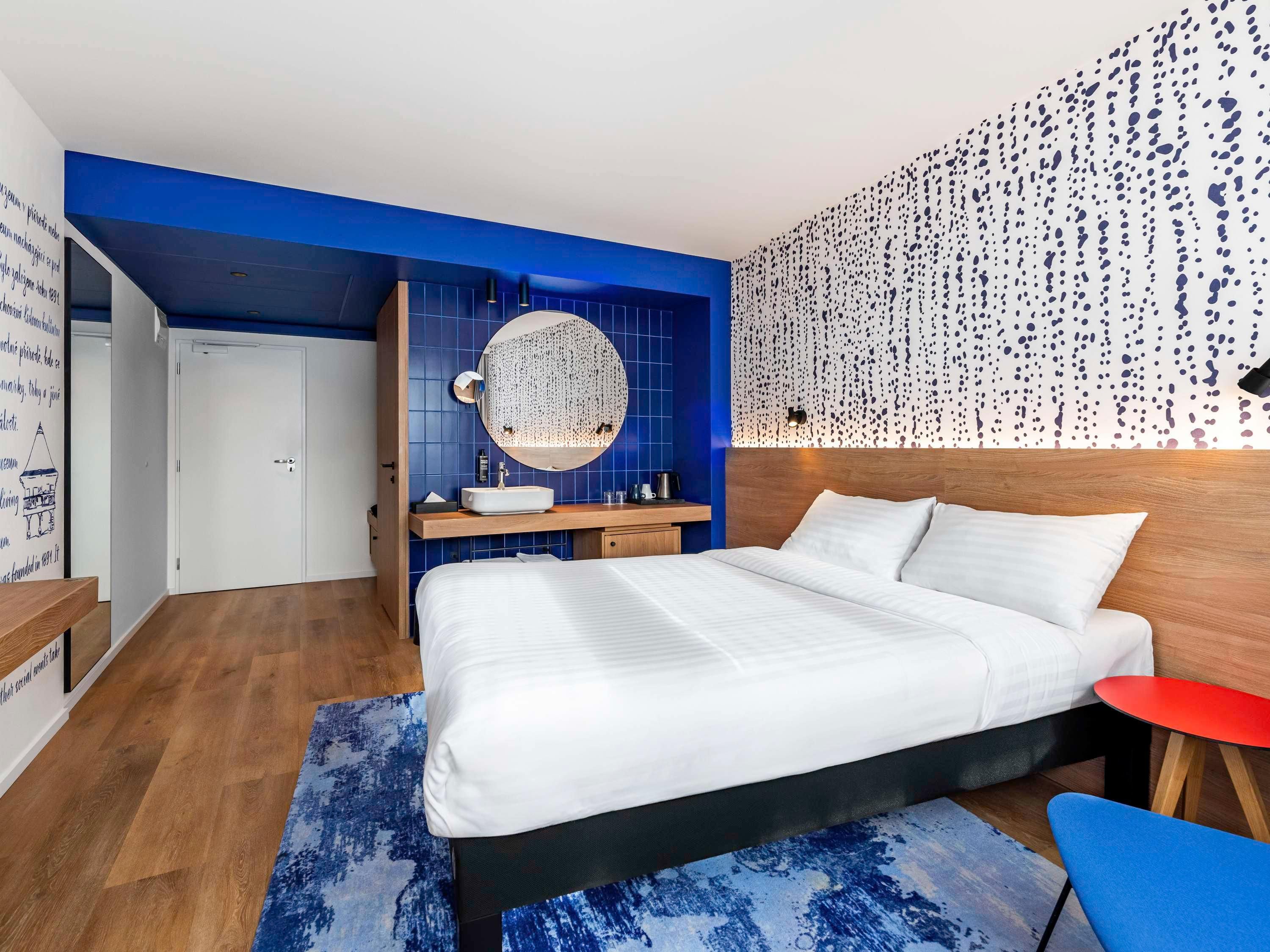 A modern bedroom with a large bed, a round mirror, a wooden nightstand, and a red side table.