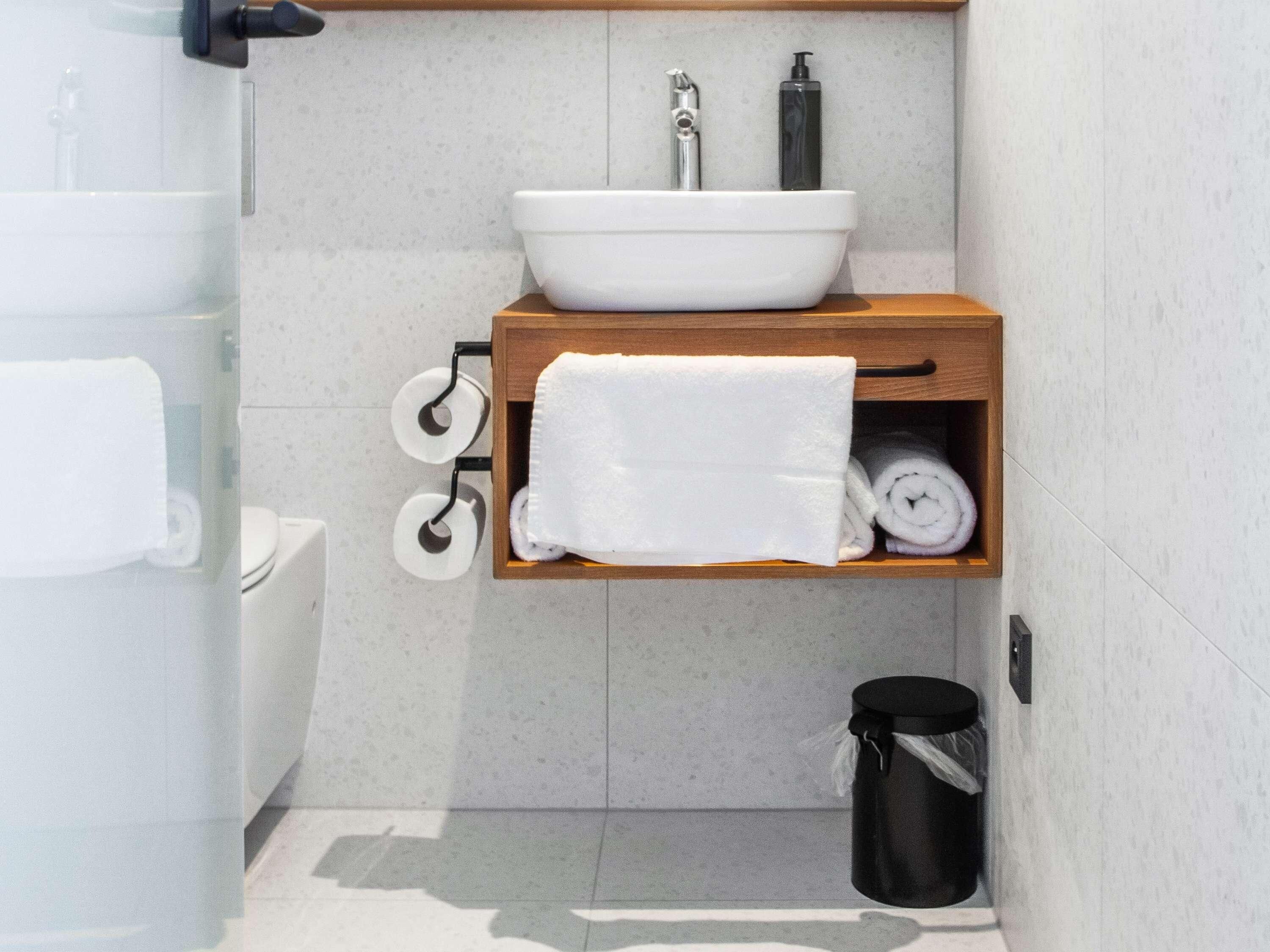 Une salle de bain avec un lavabo blanc, une étagère en bois où sont posées des serviettes, du papier toilette et une poubelle.