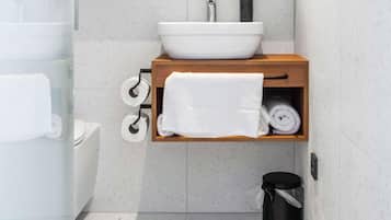 Une salle de bain avec un lavabo blanc, une étagère en bois où sont posées des serviettes, du papier toilette et une poubelle.