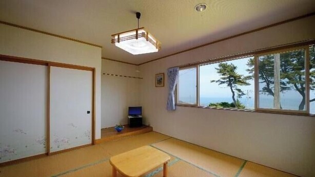 Ett traditionellt japanskt rum med tatamimattor på golvet, en skjutdörr, en tv och ett stort fönster med utsikt över träd och havet.