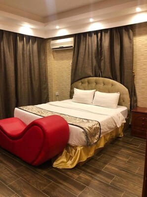 A bedroom with a tufted headboard, a bed with white linens, a red bench, and a wooden floor. - Al Wouroud Serviced Appartment (Khamis Mushait)