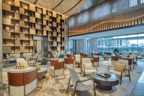 A spacious lobby with multiple seating areas, a large bookshelf, and a view of the cityscape through large windows. - Voco Xiong An (Baoding)