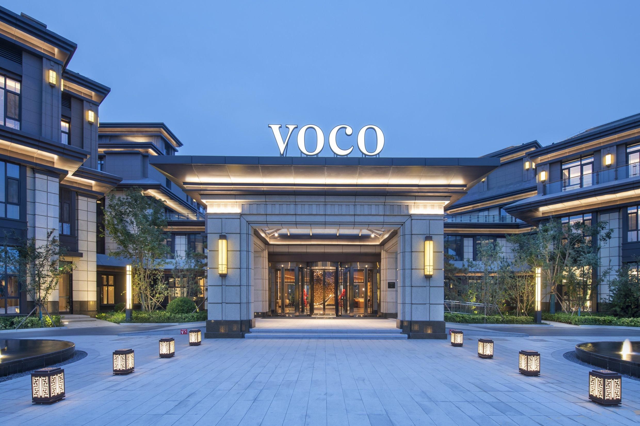 A modern hotel entrance with a prominent "VOCO" sign, a well-lit entrance, and a landscaped area with lanterns.