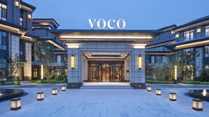 Une entrée d’hôtel moderne, avec une enseigne « VOCO » bien visible, un éclairage soigné et un espace paysager agrémenté de lanternes.