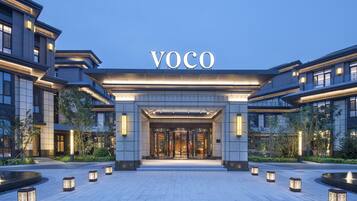A modern hotel entrance with a prominent "VOCO" sign, a well-lit entrance, and a landscaped area with lanterns.