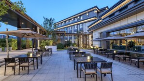 An outdoor patio area with multiple tables and chairs, some with umbrellas, and a modern building in the background. - Voco Xiong An (Baoding)