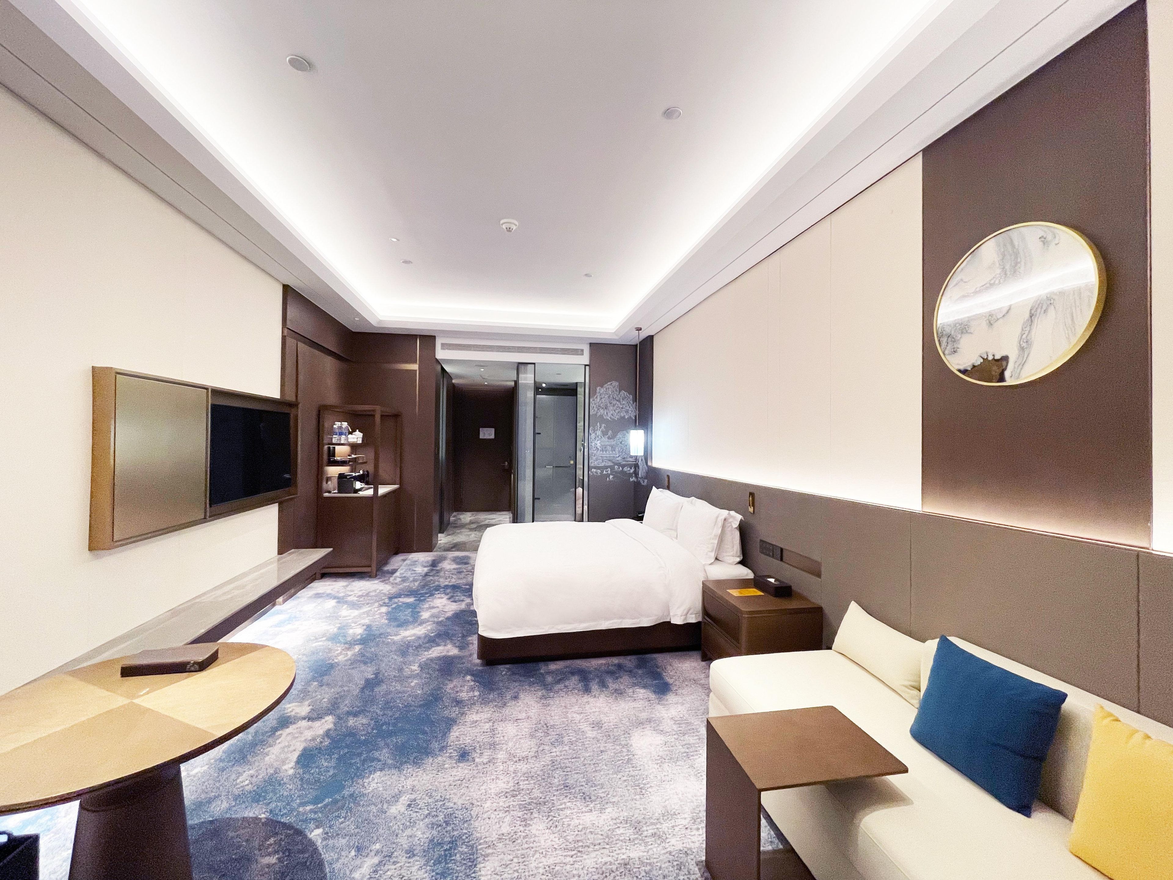 A hotel room with a large bed, a sofa, a desk, a TV, and a bathroom visible through an open door.