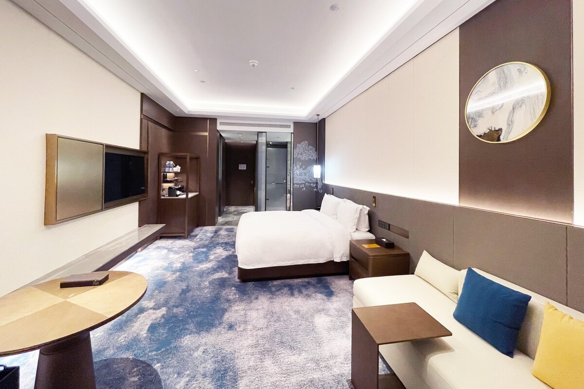 A hotel room with a large bed, a sofa, a desk, a TV, and a bathroom visible through an open door.