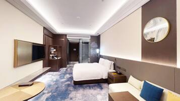 A hotel room with a large bed, a sofa, a desk, a TV, and a bathroom visible through an open door.