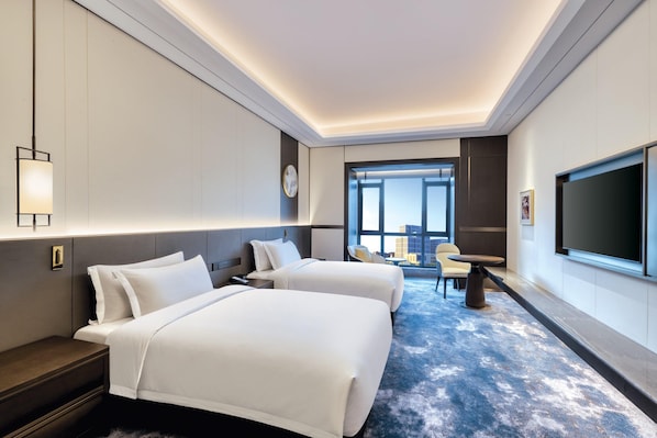 A hotel room with two beds, a desk, a chair, a TV, and a large window with a view.