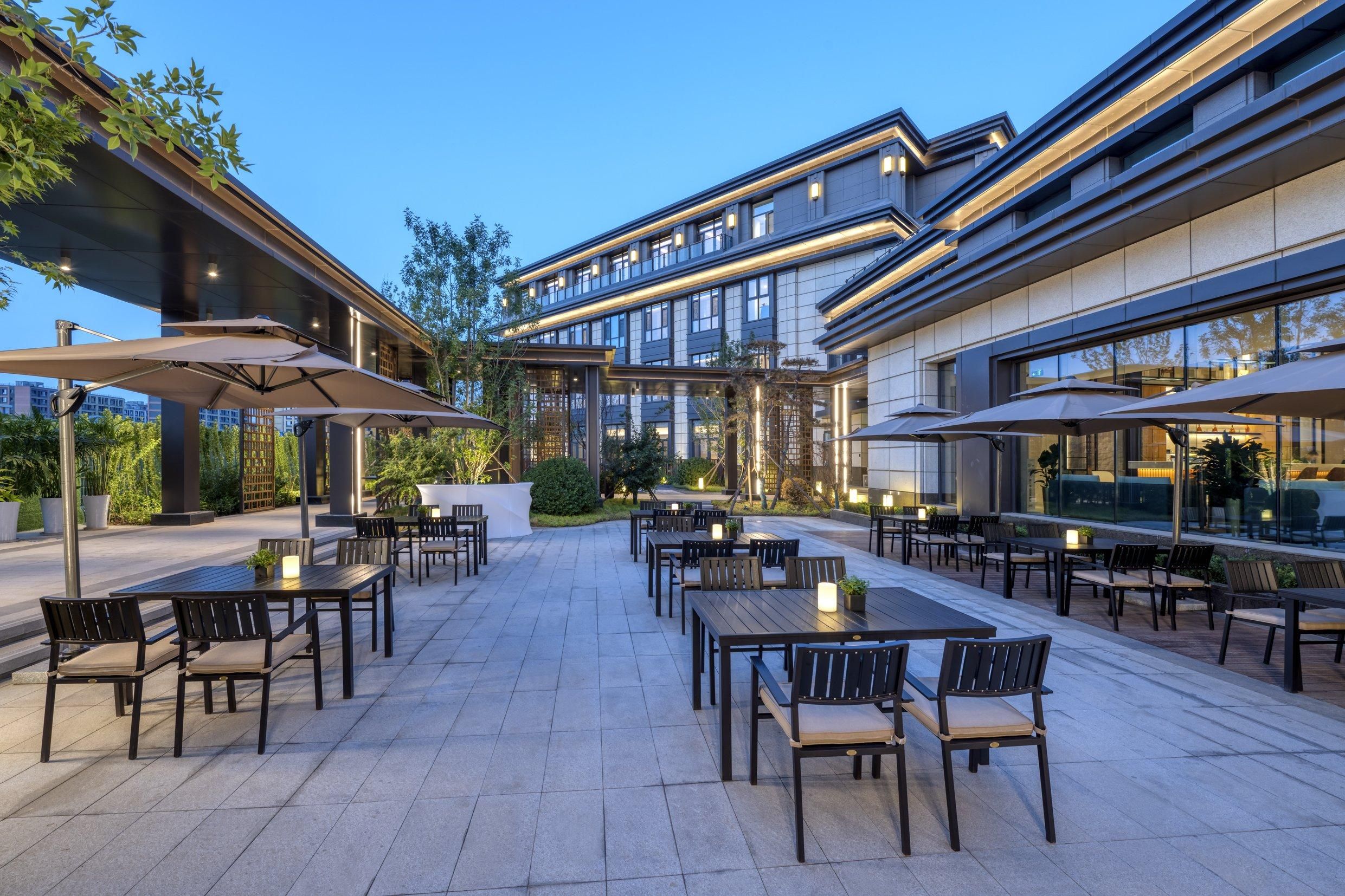 Un patio al aire libre con varias mesas y sillas, algunas con sombrillas, y un edificio moderno al fondo.