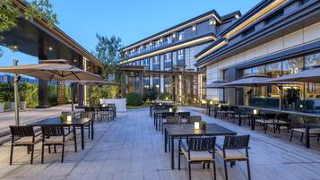 An outdoor patio area with multiple tables and chairs, some with umbrellas, and a modern building in the background.