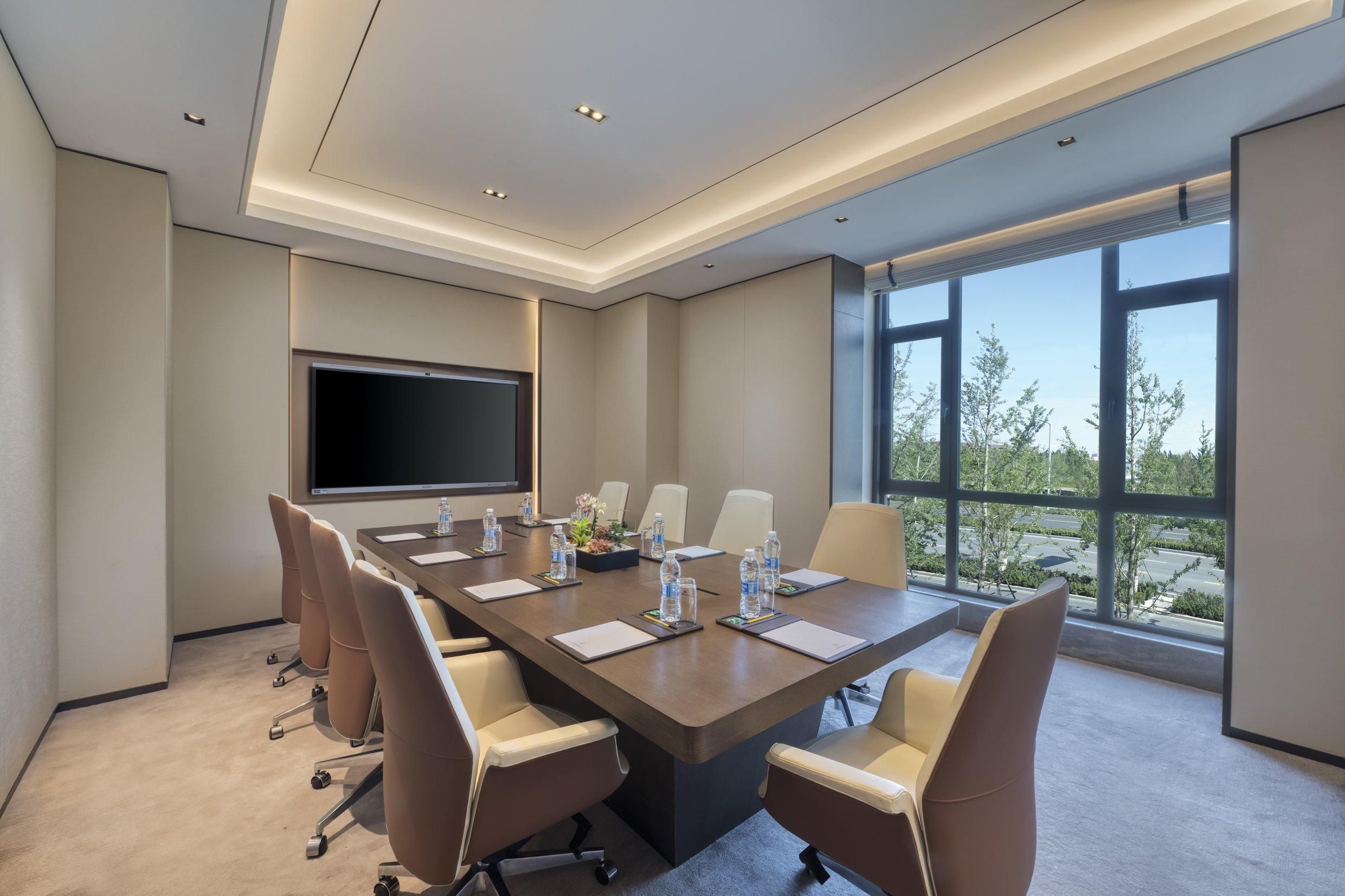 A conference room with a long table, chairs, a flat-screen TV, and large windows with a view of trees and a road.