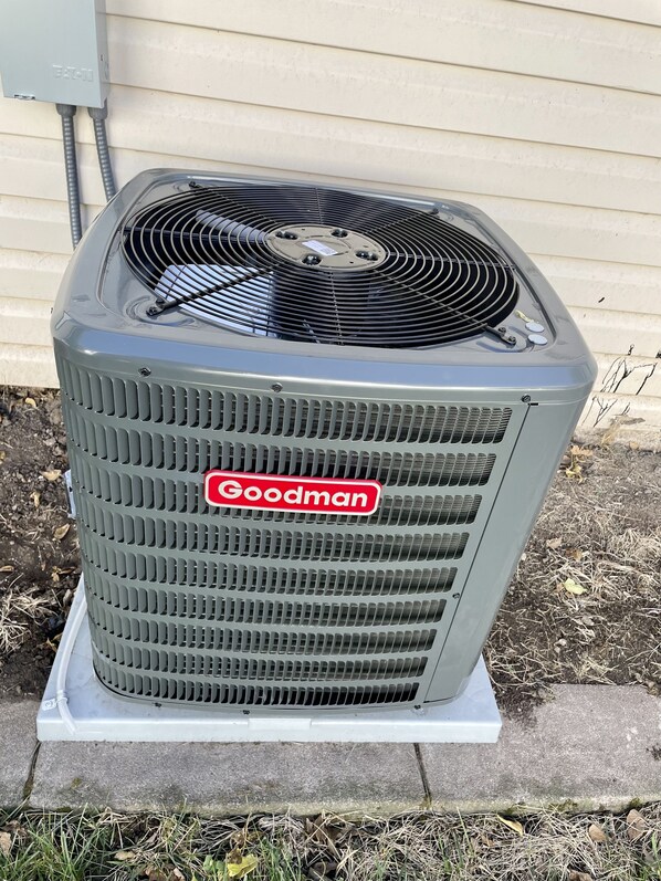 A stationary Goodman air conditioning unit mounted on a concrete base outdoors.