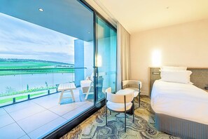 Une chambre d’hôtel dotée d’une grande porte-fenêtre coulissante donnant sur un balcon avec vue sur un lac et des champs. La chambre comprend un lit, une chaise et une petite table.