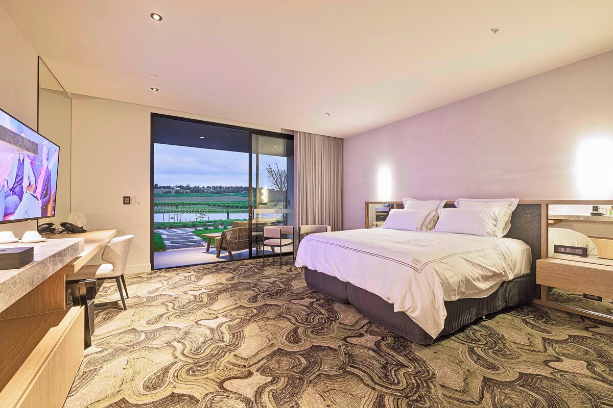 Habitación de hotel con una cama grande, un escritorio, una silla, un televisor y un balcón con vistas a un campo y una masa de agua.
