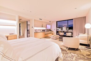A hotel room with a large bed, a desk, a chair, a sofa, a television, and a view of the city at night. - Re'em Yarra Valley (Melbourne)